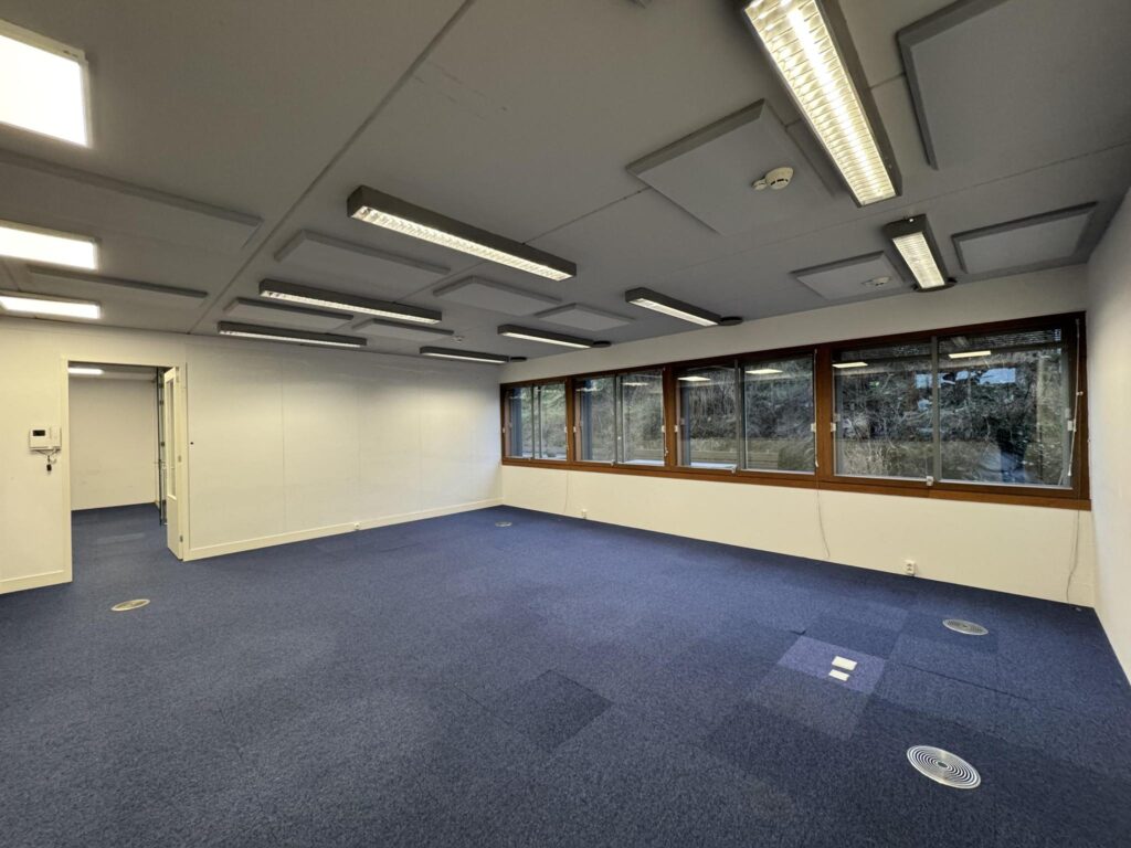 Empty office space with blue carpet flooring, fluorescent ceiling lights, and large windows at De Ruijterkade.